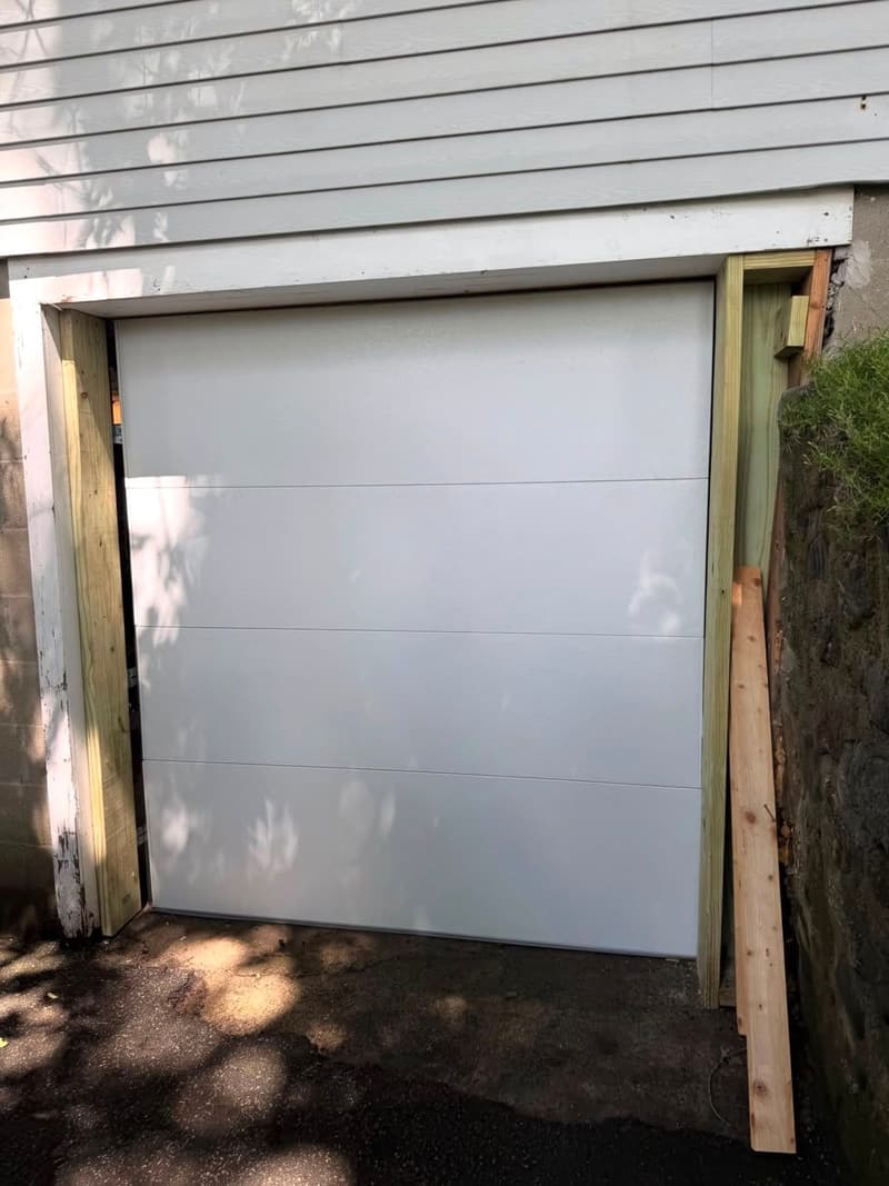 New white insulated flush-panel single garage door with fresh wood framing on the same opening