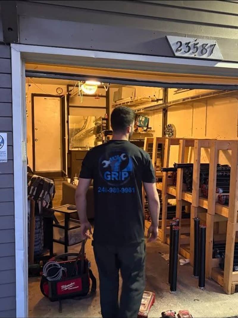 A Grip Garage technician in a branded shirt walking into a lit workshop stocked with garage door springs and parts